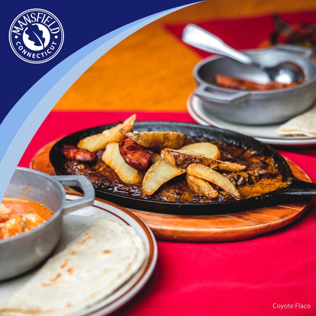 A skillet full of churrasco sits on a red table cloth, dishes to either side are obscured